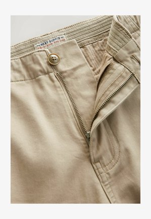 Bež hlače iz bombaža z zadrgo, gumbnim zapiranjem in elastičnim pasom. Vidno šivanje ob šivih in zadrgi.