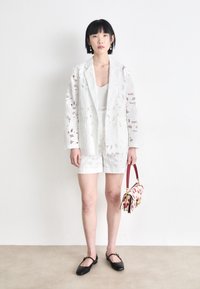 White lace blazer with floral cutouts, paired with a white camisole and shorts. Black flats and a floral handbag complete the outfit.