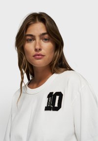 Witte katoenen t-shirt met een zwart vilt patch met het nummer "10" en de tekst "DAYS" op de linkerkant van de borst. Soepele textuur, relaxte fit.