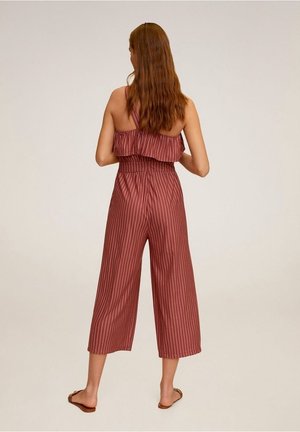 Femme aux longs cheveux portant une combinaison sans manches bordeaux avec des rayures verticales blanches, debout pieds nus sur un fond uni, de dos.