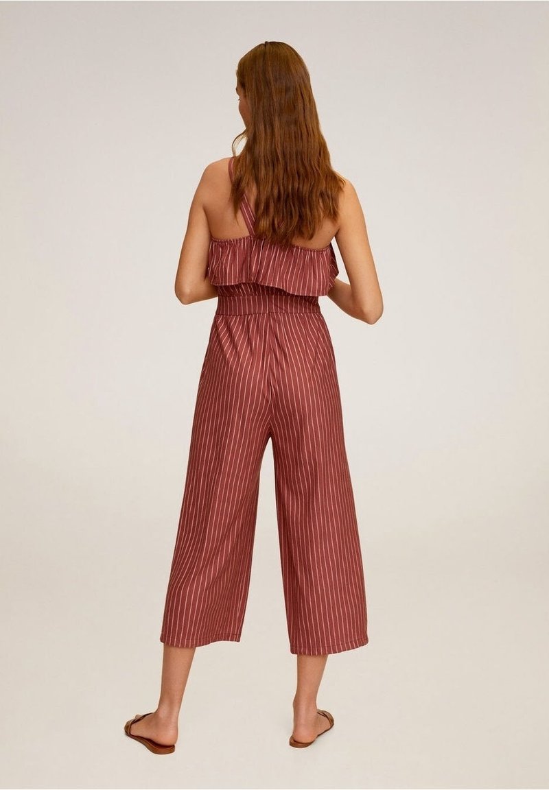 Femme aux longs cheveux portant une combinaison sans manches bordeaux avec des rayures verticales blanches, debout pieds nus sur un fond uni, de dos.