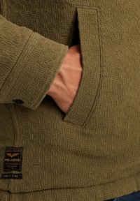 Ręka w kieszeni teksturowanej oliwkowo-zielonej kurtki z czarnym guzikiem na mankiecie i metką marki PME Legend blisko dołu.