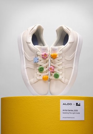 Baskets blanches avec un upper en synthétique texturé, ornées de charme en forme de fleurs colorées et de cœurs sur les lacets, présentées sur un podium jaune.