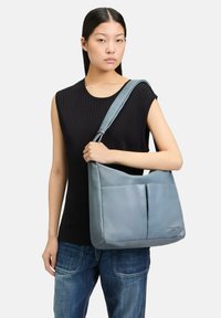 Borsa a spalla blu in pelle con una texture liscia, dotata di due tasche anteriori e di una tracolla larga, indossata da una persona in abbigliamento casual.
