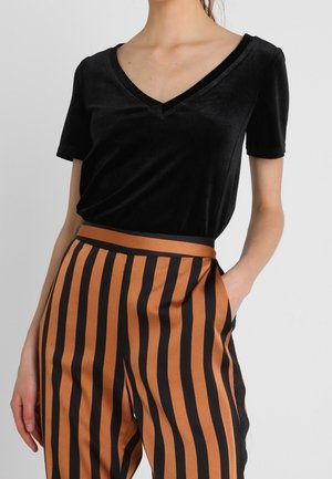 Kvinde iført en sort fløjl kortærmet V-hals top stoppet ned i højtaljede brune bukser med vertikale sorte striber.