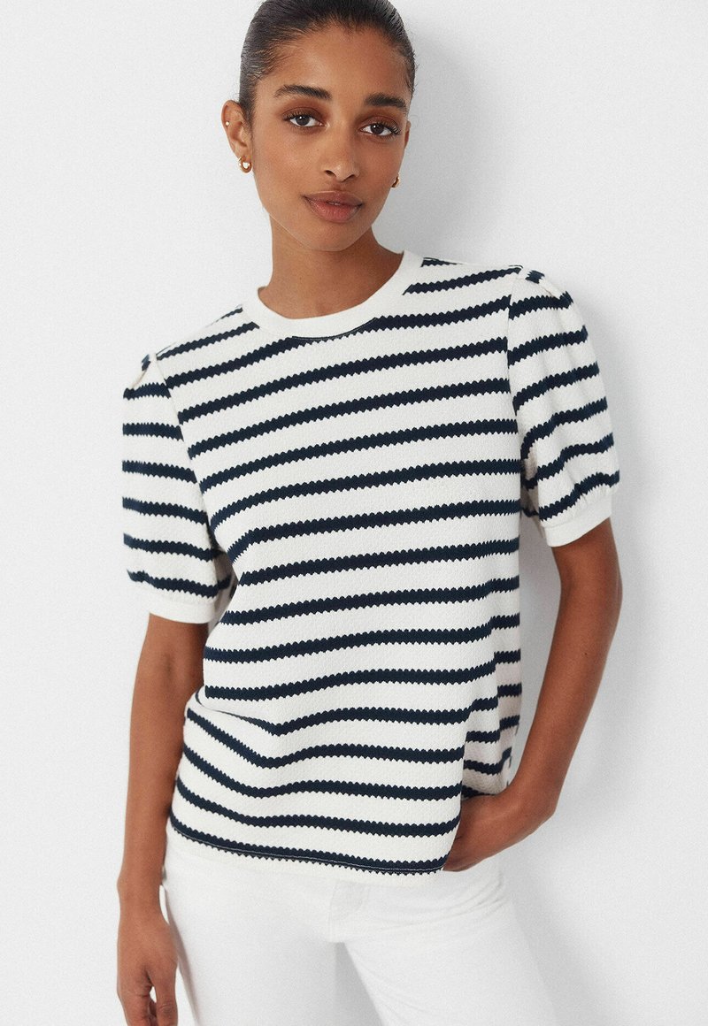 Jonge vrouw met haar in een knot, draagend gouden oorringen en een wit kortermig shirt met zwarte horizontale strepen, poserend met één hand in haar zak.