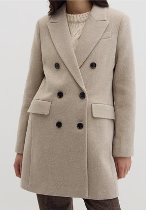 Femme portant un manteau en laine beige à double boutonnage avec des boutons noirs et des poches à rabat sur un pull en tricot crème.