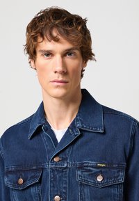 Laivastonsininen farkkutakki, jossa on kaksi etutaskua, metalliset napit ja Wrangler-logo. Ominaisuuksiin kuuluu klassinen kaulus ja pehmeä tekstuuri.