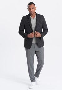 Blazer en laine gris foncé sur un polo texturé gris clair, associé à un pantalon gris clair et des baskets blanches. Le modèle se tient avec assurance, les mains jointes.