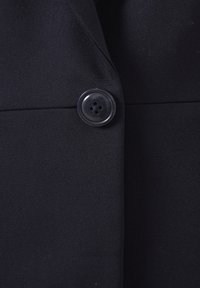 Tissu de blazer noir avec un seul grand bouton rond noir fixé à la couture centrale avant.