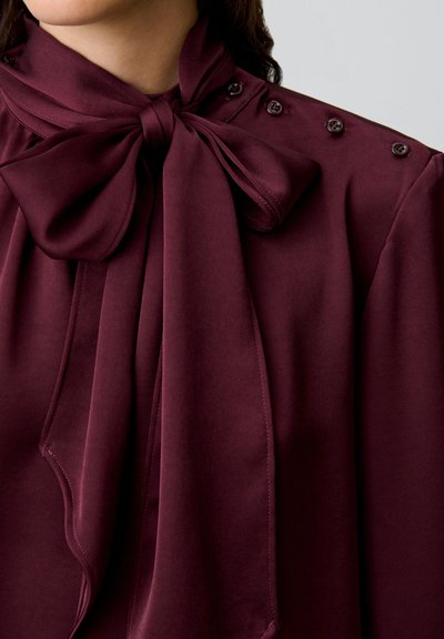 Blusa color bordeaux con un grande fiocco al collo, tessuto morbido e bottoni decorativi sulla spalla. Design con drappeggio fluido.