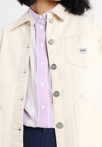 Person som bär en krämfärgad Lee-jacka över en lila och vit randig skjorta med knappar och mörkblå jeans.