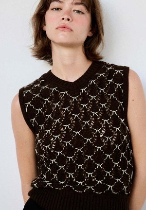 Joven con cabello corto y castaño que lleva una camiseta negra sin mangas de punto con patrones geométricos blancos sobre un fondo liso.