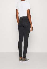 Jeans skinny negros de cintura alta, material de denim, diseño de cinco bolsillos con costuras sutiles, combinados con zapatillas bajas de color verde oliva.