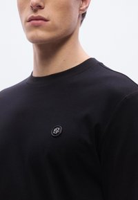 Musta puuvillainen collegepaita, jossa pyöreä pääntie, lyhyet hihat ja pieni pyöreä logopatch vasemmassa rinnassa. Sileä pinta.