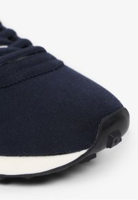 Marineblauwe sneaker met een zachte, gestructureerde bovenkant, een witte middenzool en een zwarte rubberen buitenzool met grippatronen. Veters zijn zichtbaar.