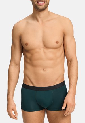 Shirtless man with a muscular build wearing dark green boxer briefs with a black waistband, standing against a plain background.