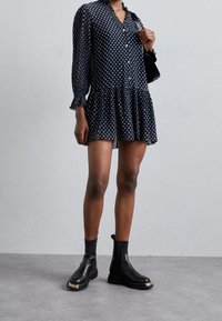 Kvinna klädd i en marinblå prickig klänning med genomskinliga ärmar och svarta ank stövlar med guldaccenter, hållande en liten svart handväska.