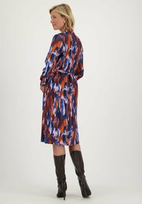 Kleurrijke patroonjurk met blauwe, oranje en paarse tinten, een getailleerde taille en lange mouwen. Knie-lang, gecombineerd met zwarte knie-hoge laarzen.