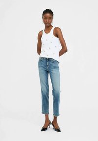 Bela majica brez rokavov z okrasnimi dodatki, združena s svetlo modrimi kavbojkami s širokimi hlačnicami in črnimi čevlji z zašiljeno konico.