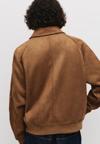 Veste en suédine marron avec une coupe décontractée, dotée d'un col, de poignets côtelés et d'une texture lisse. Détail de couture à l'arrière visible.