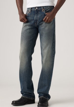 Mand iført falmede blå jeans med lige ben, sorte sko og en hvid skjorte, hænder hvilende nær de forreste lommer.