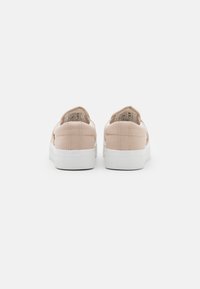 Beige canvas slip-on sneakers with a textured white rubber sole, featuring symmetrical stitching and a logo detail on the back.