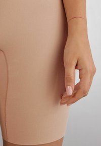 Nude-kleurige aansluitende shorts met een gladde textuur en een lichte glans, met een gestikte naaddetail aan de zijkant en een hand die voorzichtig de stof aanraakt.