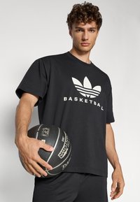 Svart bomullst-shirt med vit text "BASKETBALL" och logotyp, med korta ärmar och avslappnad passform; person som håller en basketboll.