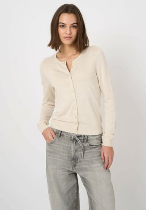 Jeune femme aux cheveux bruns mi-longs portant un cardigan beige boutonné et un jean gris clair, debout avec une main dans la poche.