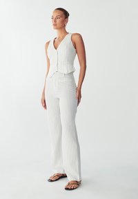 Bílá vestu s proužky a odpovídajícími širokými kalhotami. Tkanina vypadá hladce s mírným leskem. Vesta má peplum detail a knoflíky.