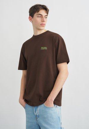 Junger Mann mit kurzen braunen Haaren, der ein braunes T-Shirt mit grünem Schriftzug "the new originals" und hellblaue Jeans trägt, die Hände in den Taschen.