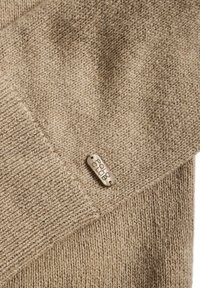 Tissu tricoté beige avec un motif texturé, doté d'une étiquette argentée gravée "POLO CLUB" attachée au bord.