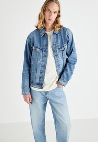 Jachetă din denim de un albastru deschis, cu închidere cu butoane, buzunare cu patch-uri și o croială lejeră. Purtați peste o cămașă de culoare crem și blugi de un albastru deschis.