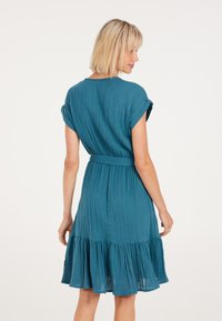 Een teal, knie-long jurk met een gestructureerde stof, korte mouwen en een brede, gerimpelde zoom. Inclusief een bijpassende riem in de taille.