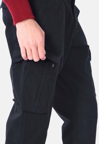 Pantaloni cargo neri con tasca laterale, tessuto robusto e cuciture a contrasto. La tasca ha una chiusura con bottone a pressione.