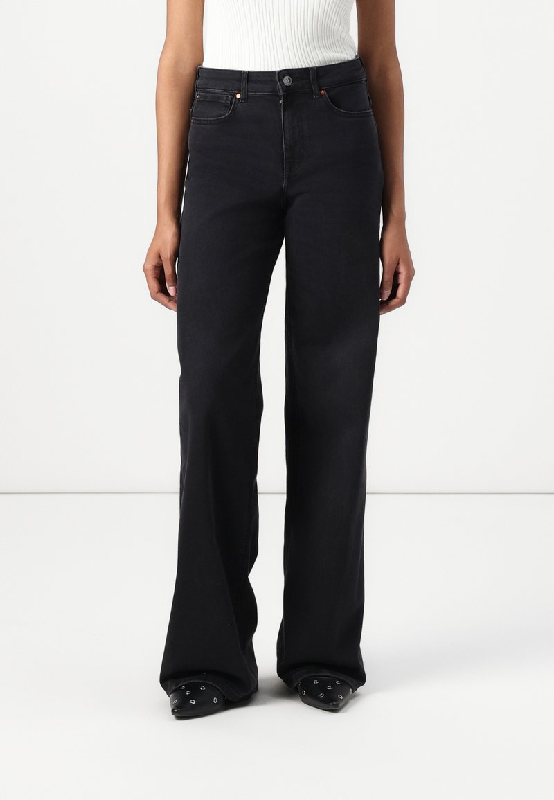 Nainen, joka on pukeutunut korkeavyötäröisiin mustiin leveälahkeisiin farkkuihin, yhdistettynä mustiin terävään kenkään, jossa on hopeiset silmukat, seisoo valkoista taustaa vasten.