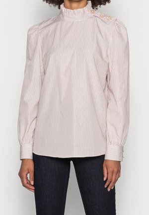 Kobieta ubrana w bluzkę z długim rękawem i wysokim kołnierzem z cienkimi pionowymi paskami i perłowymi guzikami na ramieniu, w połączeniu z ciemnymi dżinsami.