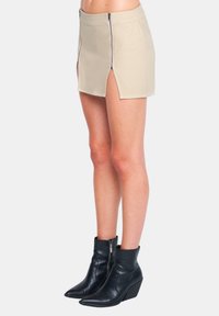 Beige Denim-Minirock mit vorderen Reißverschlüssen und seitlichen Schlitzen, kombiniert mit schwarzen Leder-Stiefeletten mit Keilabsatz.