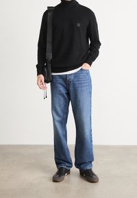Pull à col roulé noir en maille avec un petit emblème carré, associé à un jean bleu ample et des baskets noires avec des semelles marron.