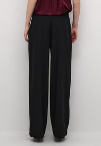 Pantaloni neri a gamba larga in tessuto liscio, con vita alta e linee pulite, abbinati a una maglietta rosso scuro sullo sfondo.