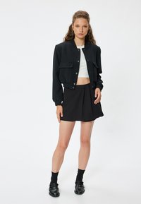 Giacca bomber nera con tasche frontali, taglio corto, abbinata a una gonna plissè nera. Il modello indossa una maglietta bianca e scarpe nere.