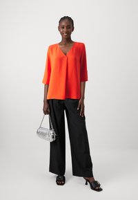 Top arancione brillante a V con maniche a tre quarti; abbinato a pantaloni neri a gamba larga. Borsa argento con tracolla a catena; décolleté neri aperti.