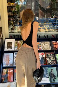 Schwarzes, rückenfreies Oberteil mit einem gedrehten Design, kombiniert mit hellbeigen, lockeren Hosen. Die Figur hält eine geflochtene schwarze Tasche. Im Hintergrund sind Magazine.