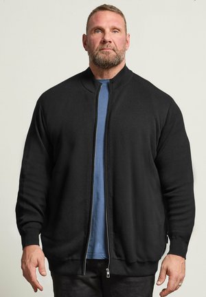 Brodaty mężczyzna w czarnej kurtce zapinanej na zamek błyskawiczny, na niebieskiej koszuli, stojący na jednolitym jasnym tle, zwrócony przodem.