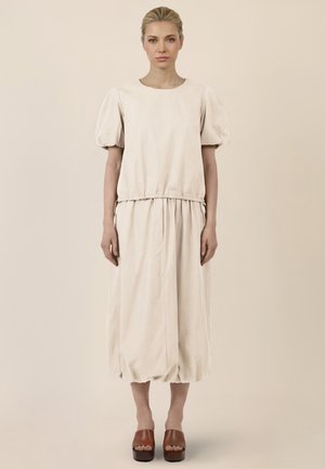 Frau mit blondem Haar, die ein lockeres, cremefarbenes Kleid mit Puffärmeln und braune Plateausandalen trägt, steht vor einem schlichten Hintergrund.