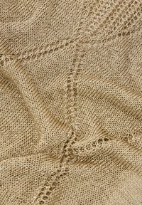 Nahaufnahme eines beigefarbenen Strickstoffs, der detaillierte, ineinander verflochtene und offene Maschenmuster zeigt, die strukturierte Falten erzeugen.