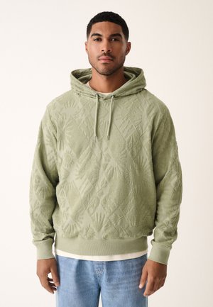 Sudadera con capucha verde hecha de tela texturizada con patrones de manos y sol. Cuenta con capucha ajustable con cordón y puños acanalados. Se lleva sobre una camiseta blanca.