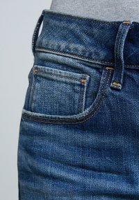 Blauwe denim jeans met een aansluitende tailleband, met een achterzak en contrasterend oranje stiksel. De stof heeft een lichte textuur en vervaging.