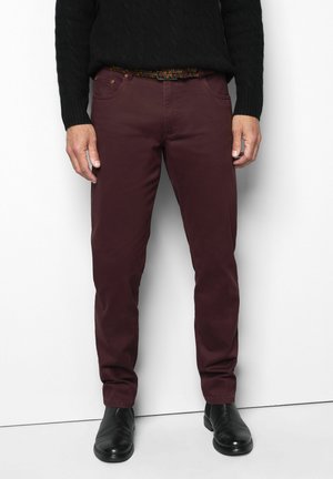 Mann trägt dunkelrote Slim-Fit-Hose, schwarze Lederschuhe, schwarzen Kabelstrickpullover und bunten gewebten Gürtel, steht vor weißem Hintergrund.